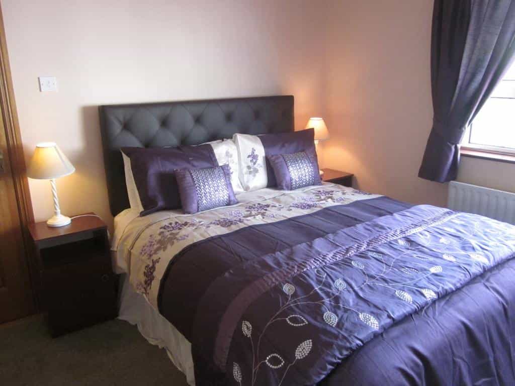 Bedroom with purple bedding, tufted headboard, bedside lamps, and dark curtains near a window.