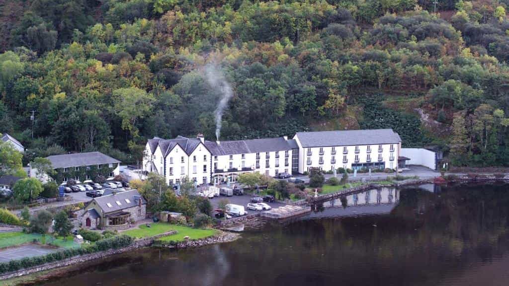 White hotel building by dark water with smoke rising, backed by dense green hillside.