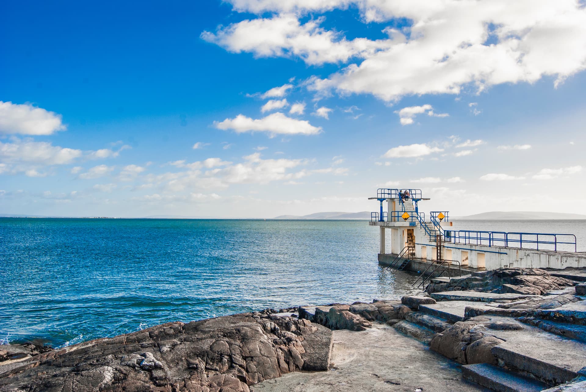 Diving platform overlooking Galway Bay with rocky foreground and blue sky.