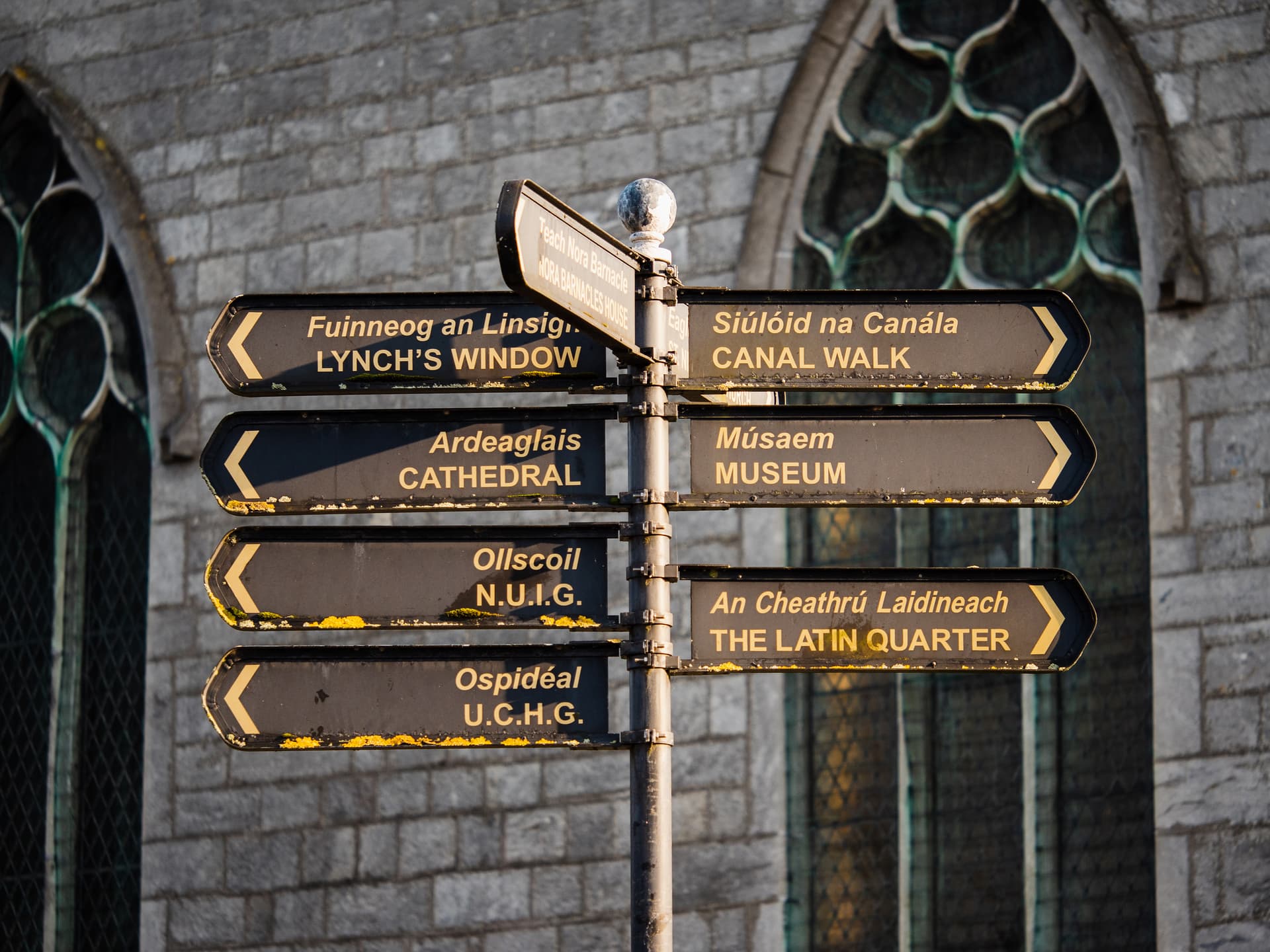 Bilingual directional signpost pointing to Cathedral and Latin Quarter against stone building in Galway.