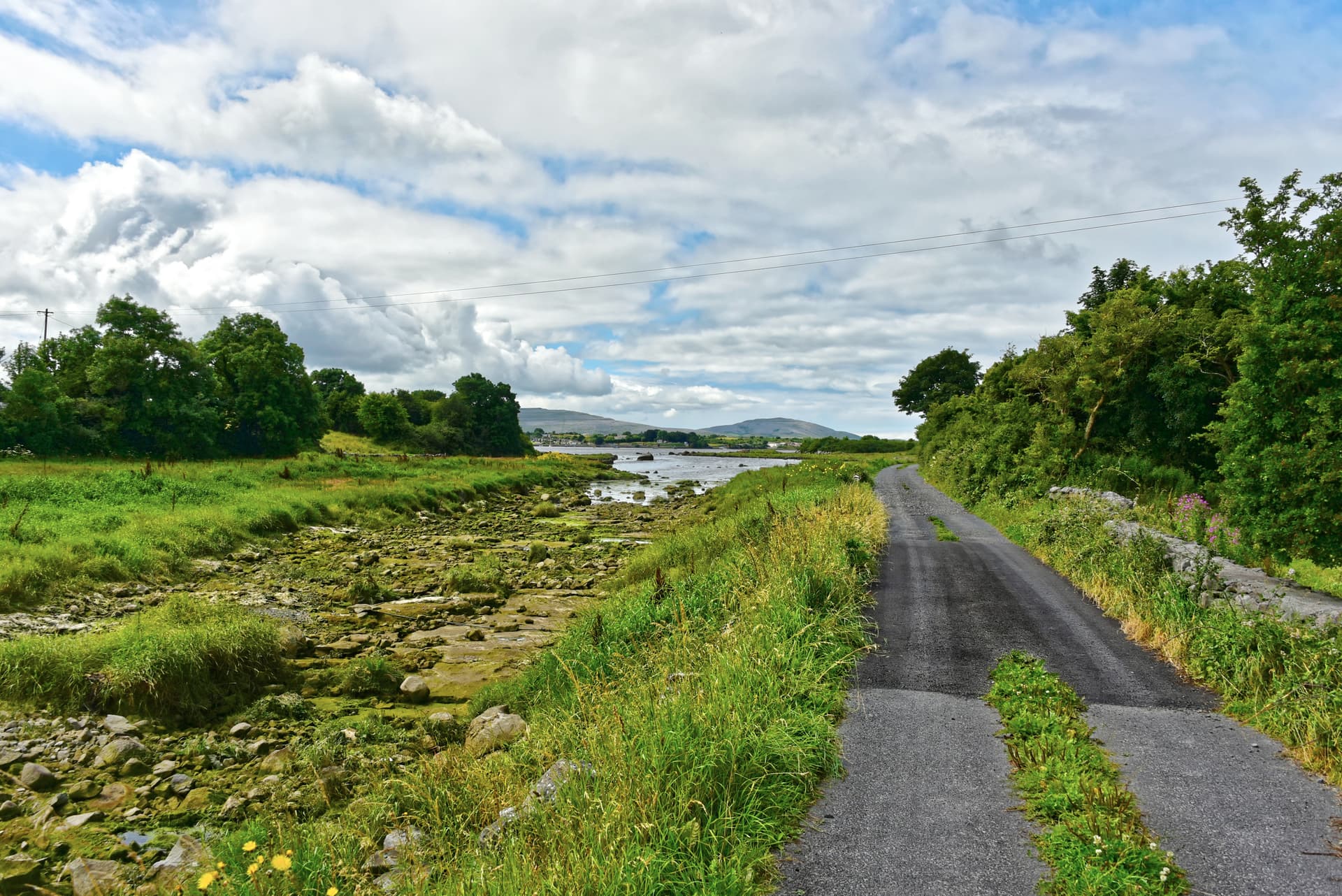 Narrow country road beside a rocky, grassy inlet under a cloudy sky, with mountains in the distance.
