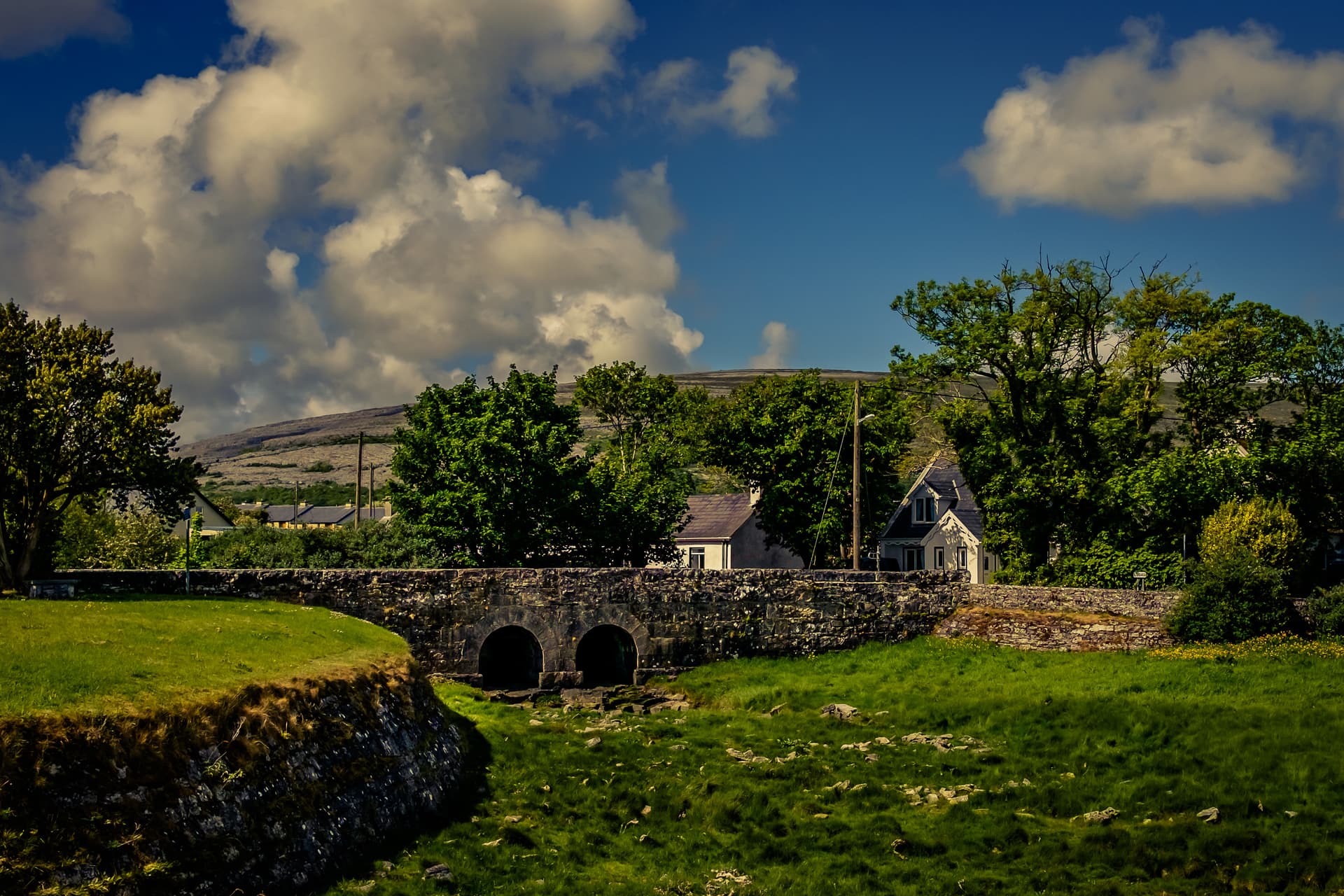 Stone arch bridge over a stream with green fields, houses, and a barren hill under a cloudy blue sky in Ballyvaughan.