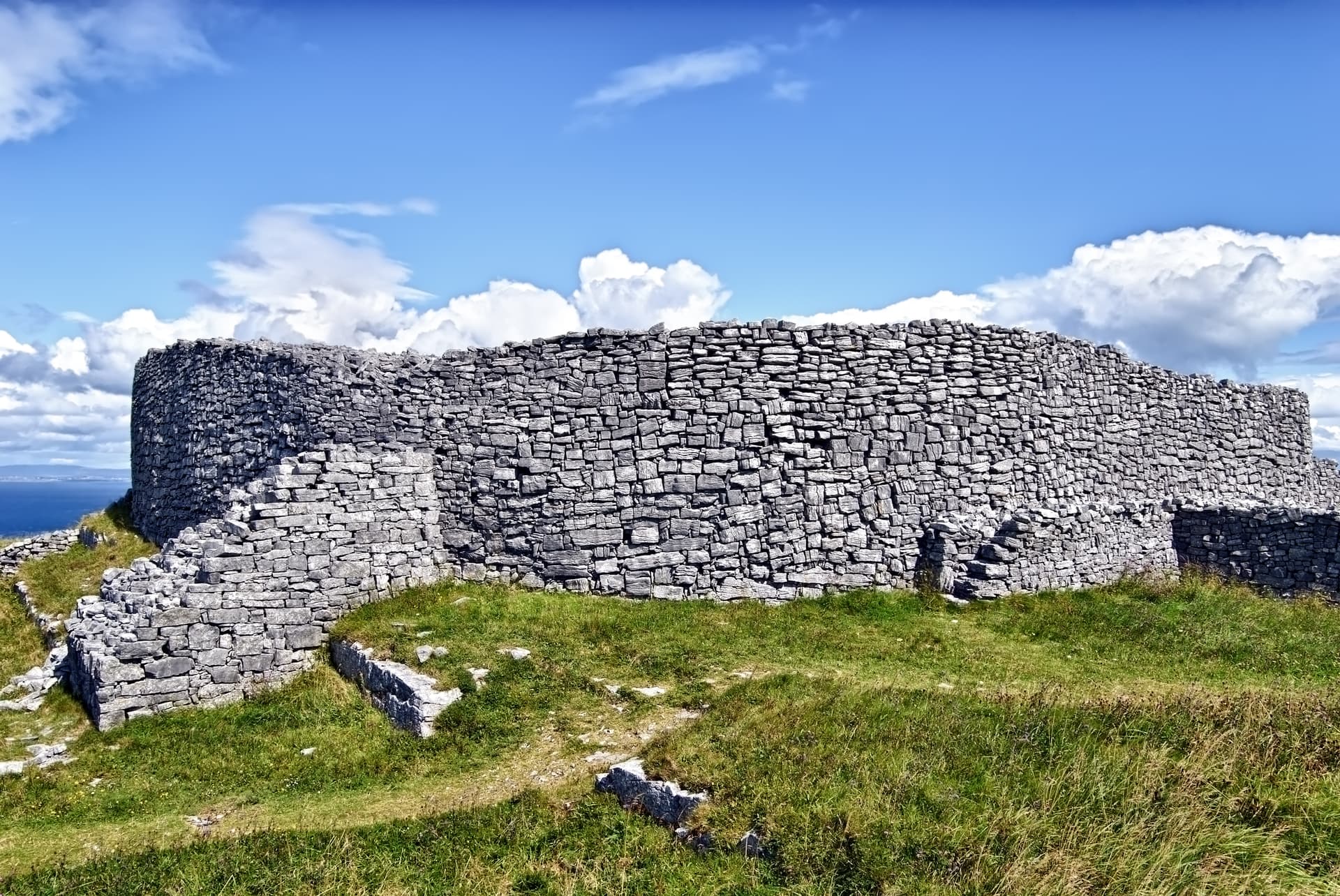Stone fort ruins on grassy hill overlooking the sea under a blue, cloudy sky at Dun Eochla, Inishmore.