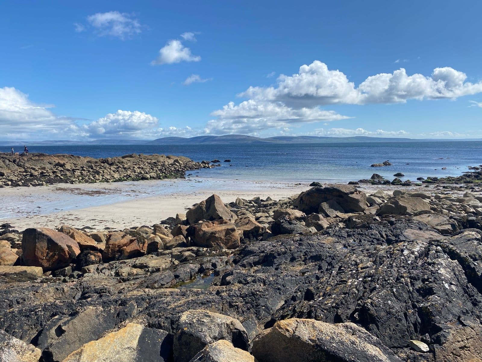 Rocky coastline with sandy beach, blue sea, and distant mountains under a cloudy sky in Spiddal.