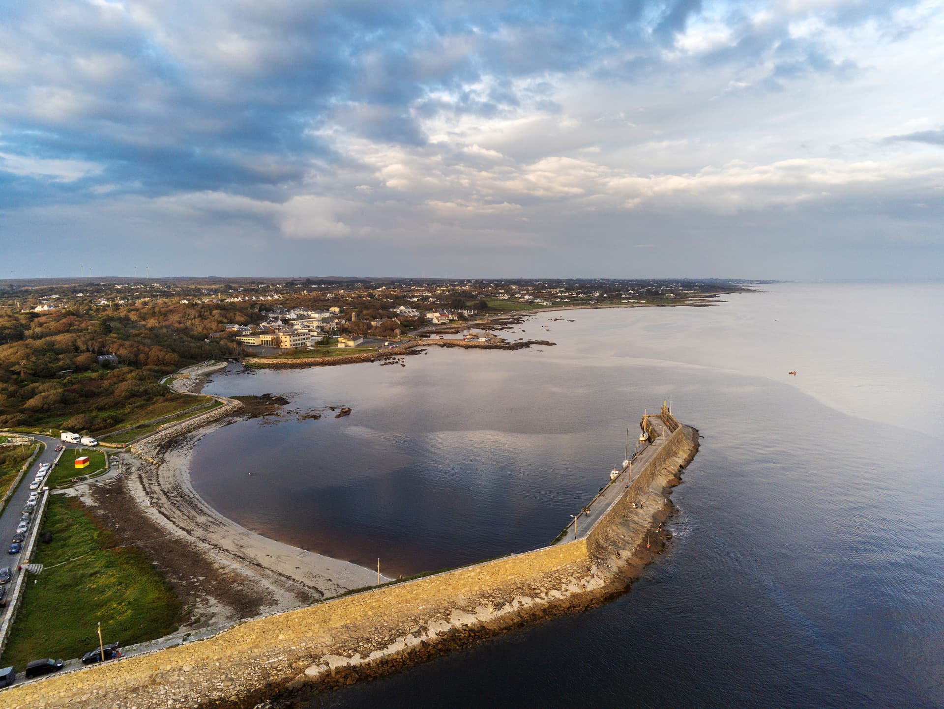 Stone pier extending into dark water near a curved beach and coastal town, Spiddal.