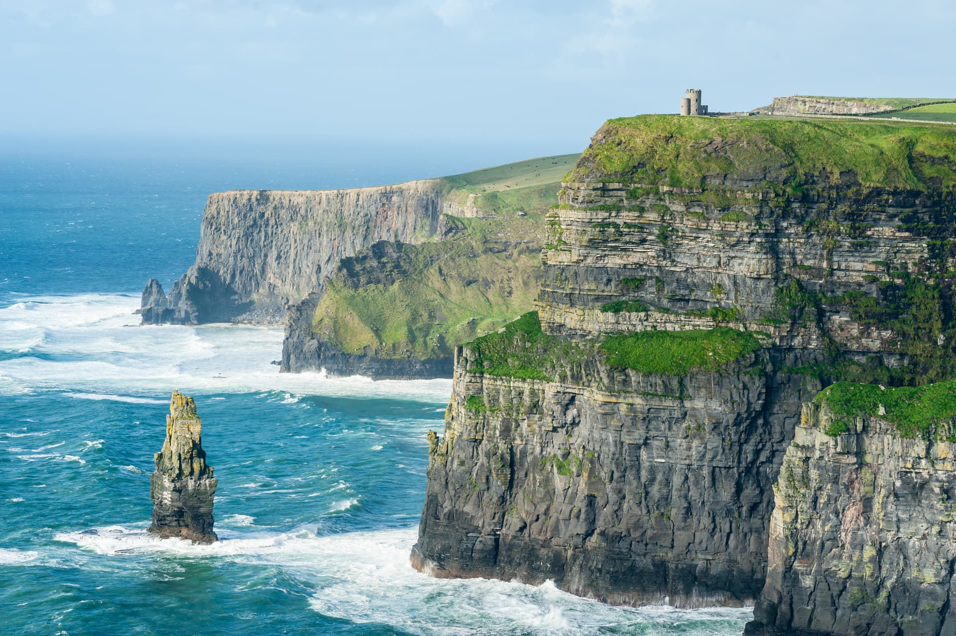Cliffs of Moher with O'Brien's Tower on grassy clifftop overlooking rough blue sea.