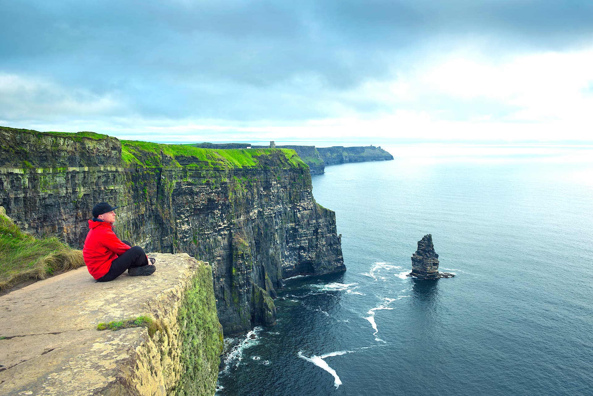 Person in red jacket sitting on edge of Cliffs of Moher overlooking sea stack and ocean