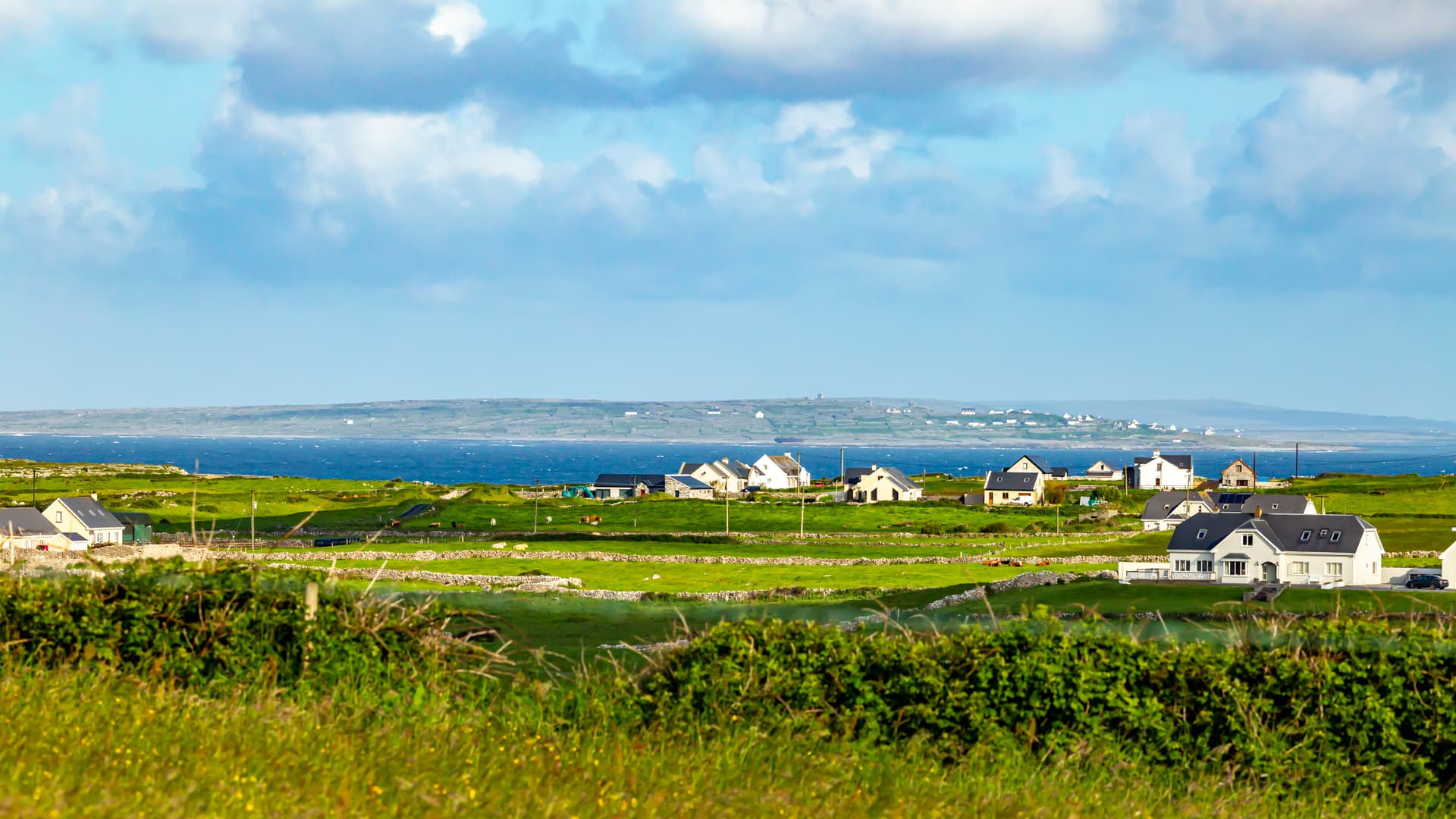 Green fields and white houses near the sea with a distant, hazy coastline under a blue sky.