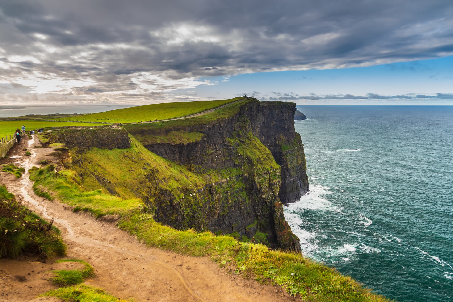 Hikers on a dirt path along the grassy Cliffs of Moher overlooking the dark green sea.