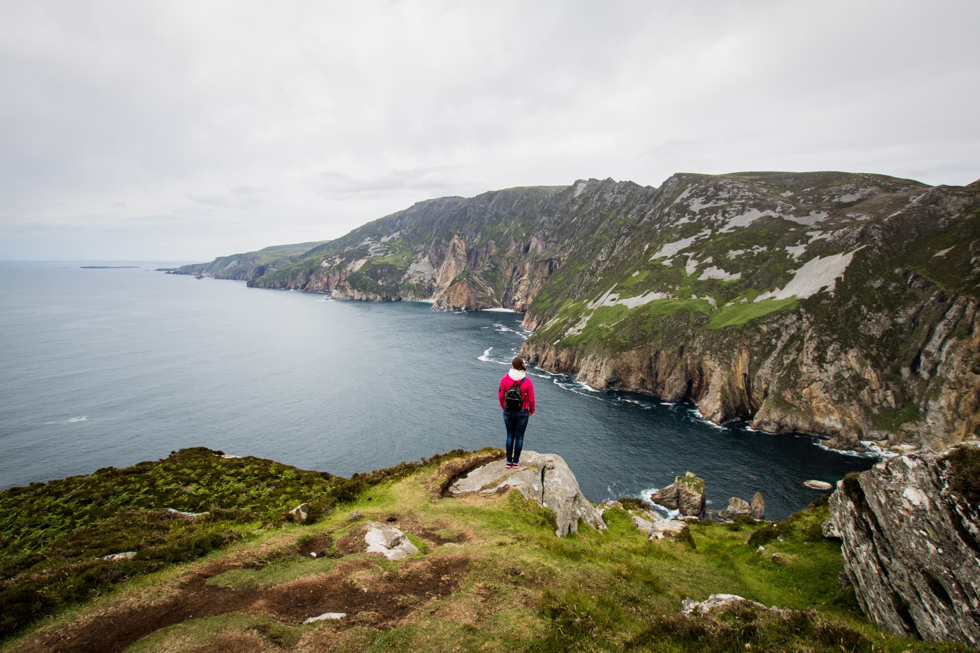 Hiker viewing dramatic sea cliffs and dark ocean water under an overcast sky, Sliab Liagh.