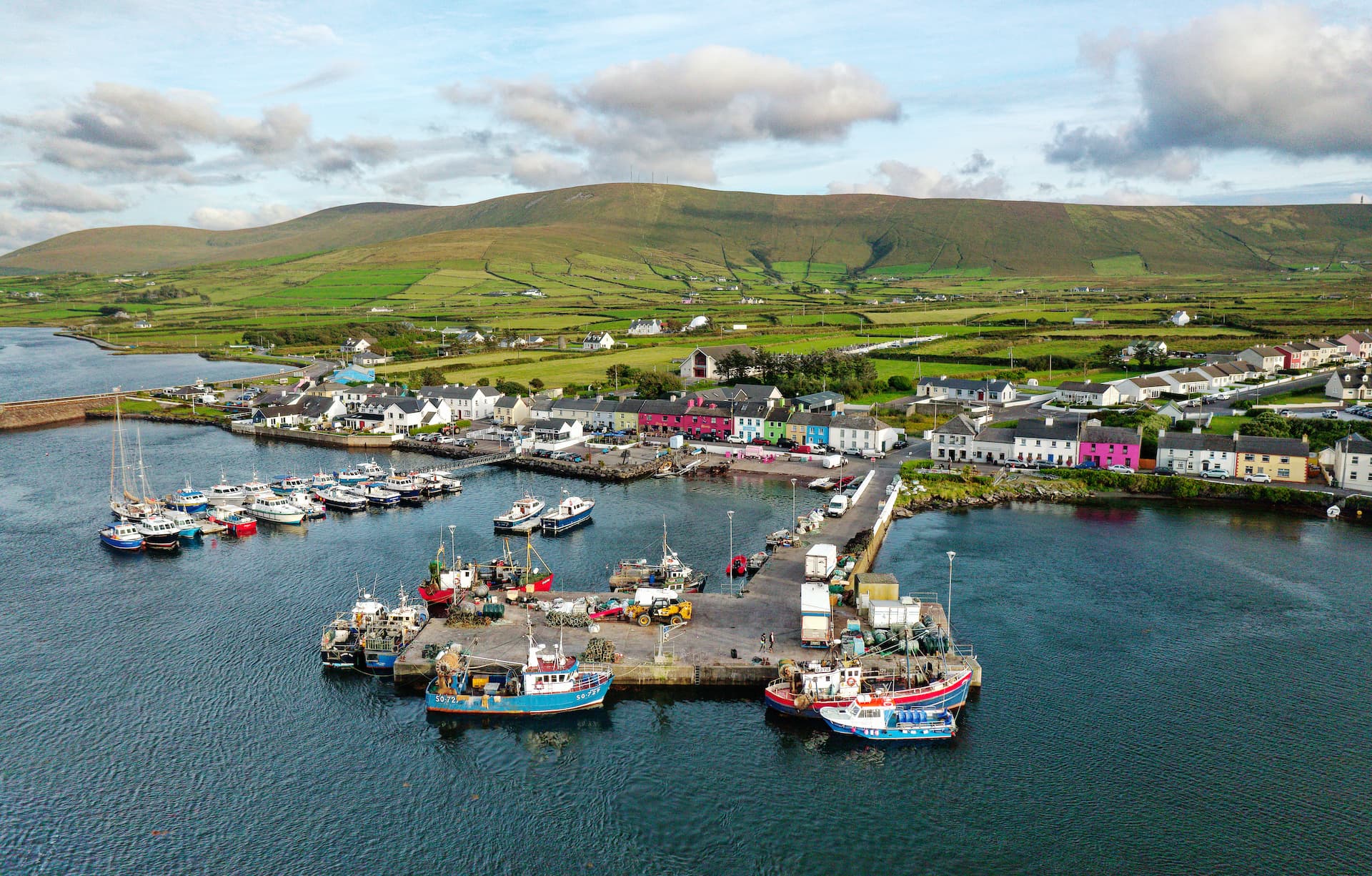 Fishing boats docked in Portmagee harbor with colorful village buildings and green hills.