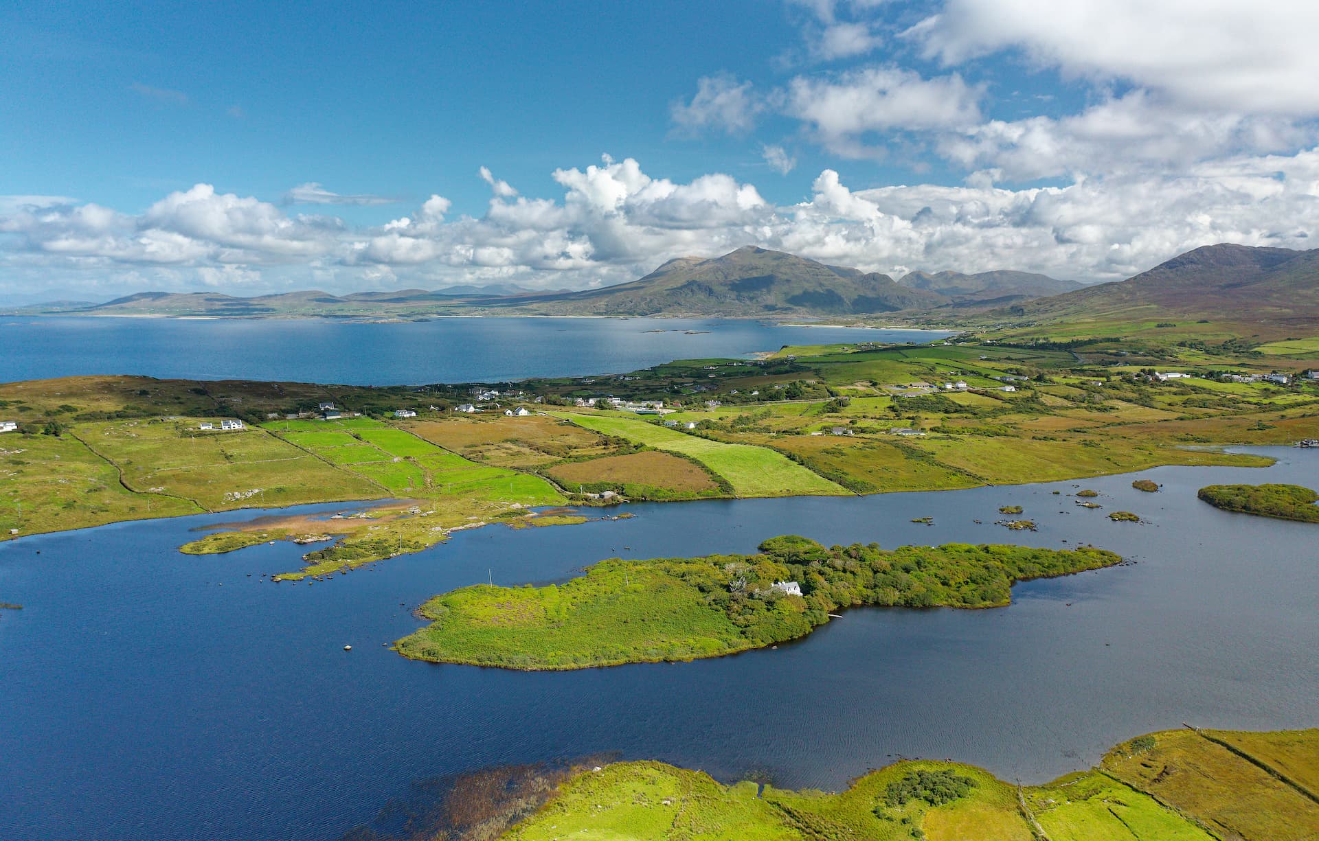 Killary Harbour inlet with green fields, mountains, and scattered white houses under a cloudy blue sky.