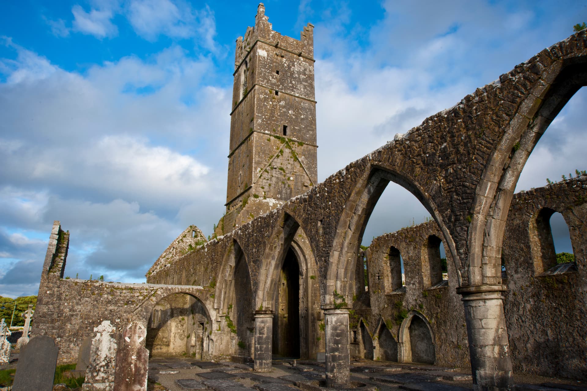 Ruins of Claregalway Friary with stone tower and arches under a blue, cloudy sky.