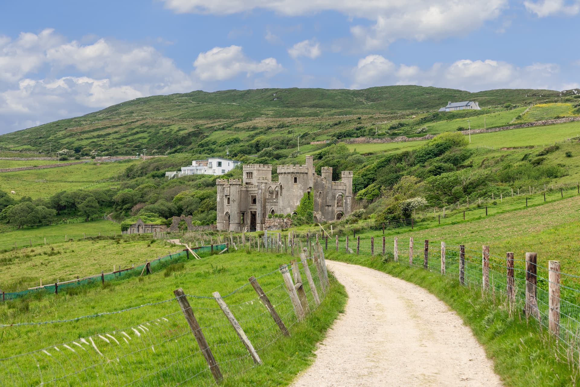 Dirt road leading to Clifden Castle ruins in green Connemara landscape under a cloudy sky.