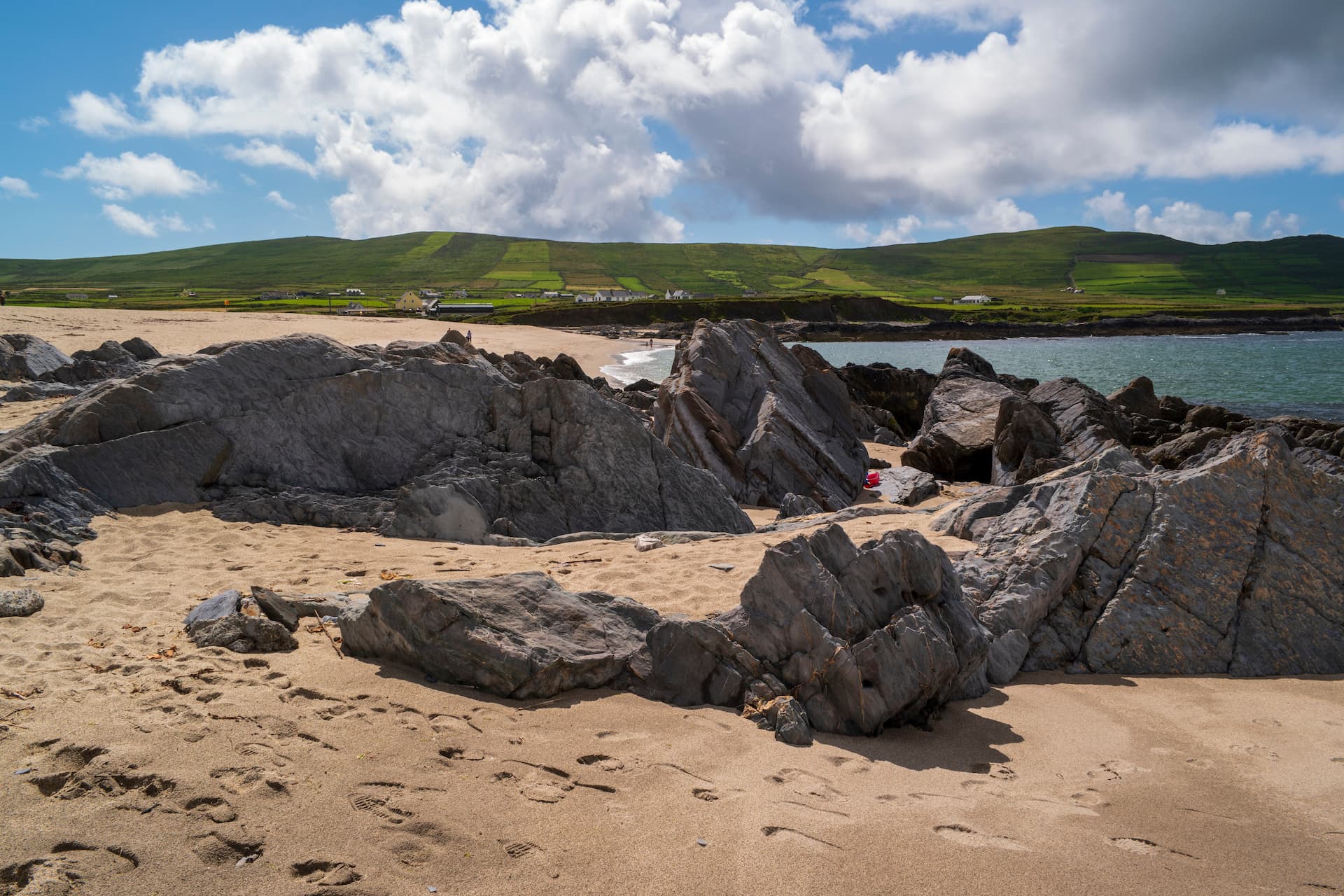 Rocky foreground on Ballydonegan Beach with green hills and blue sky overhead.