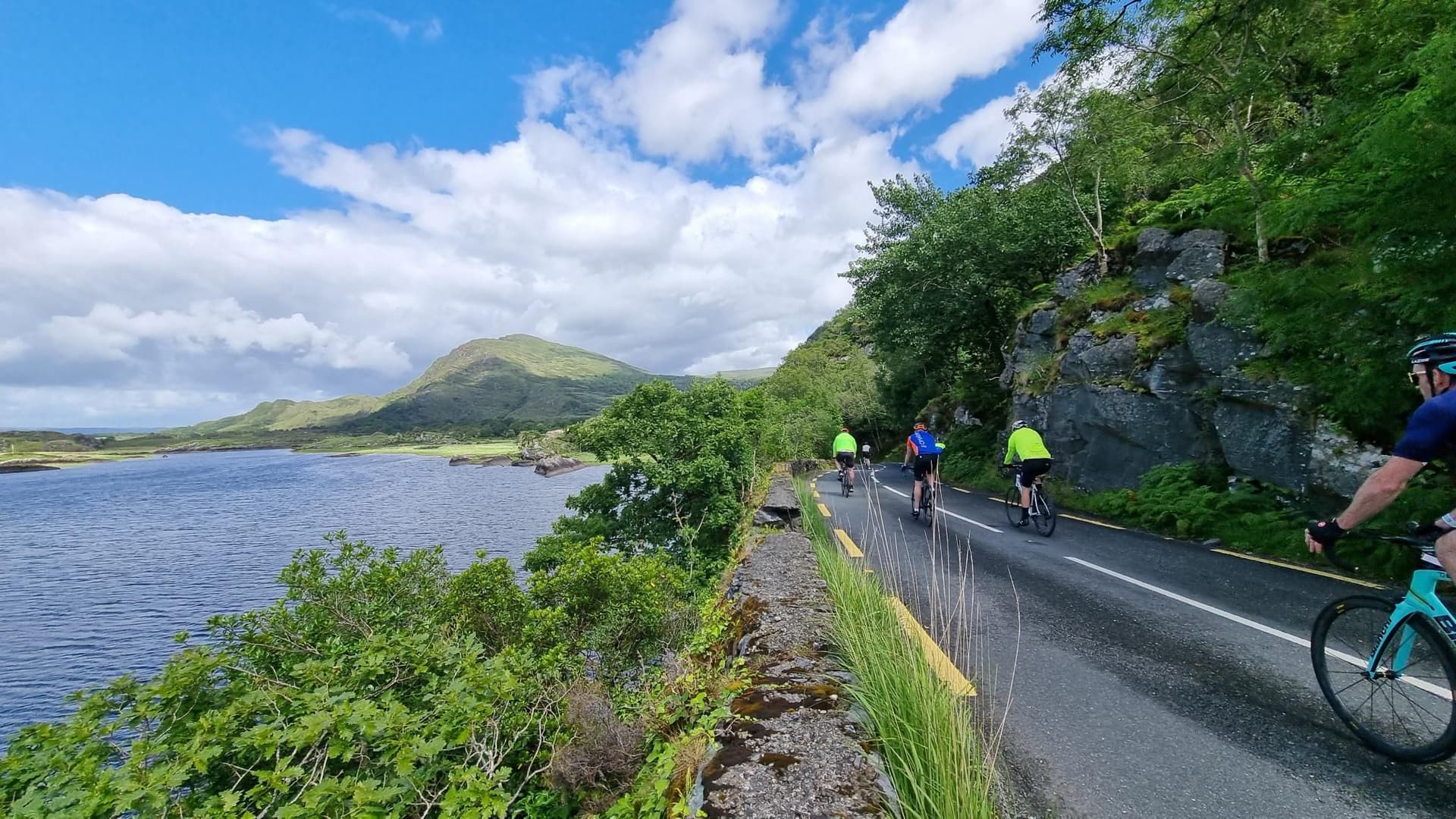 Cyclists riding along a lakeside road with green mountains under a cloudy blue sky in Killarney National Park.
