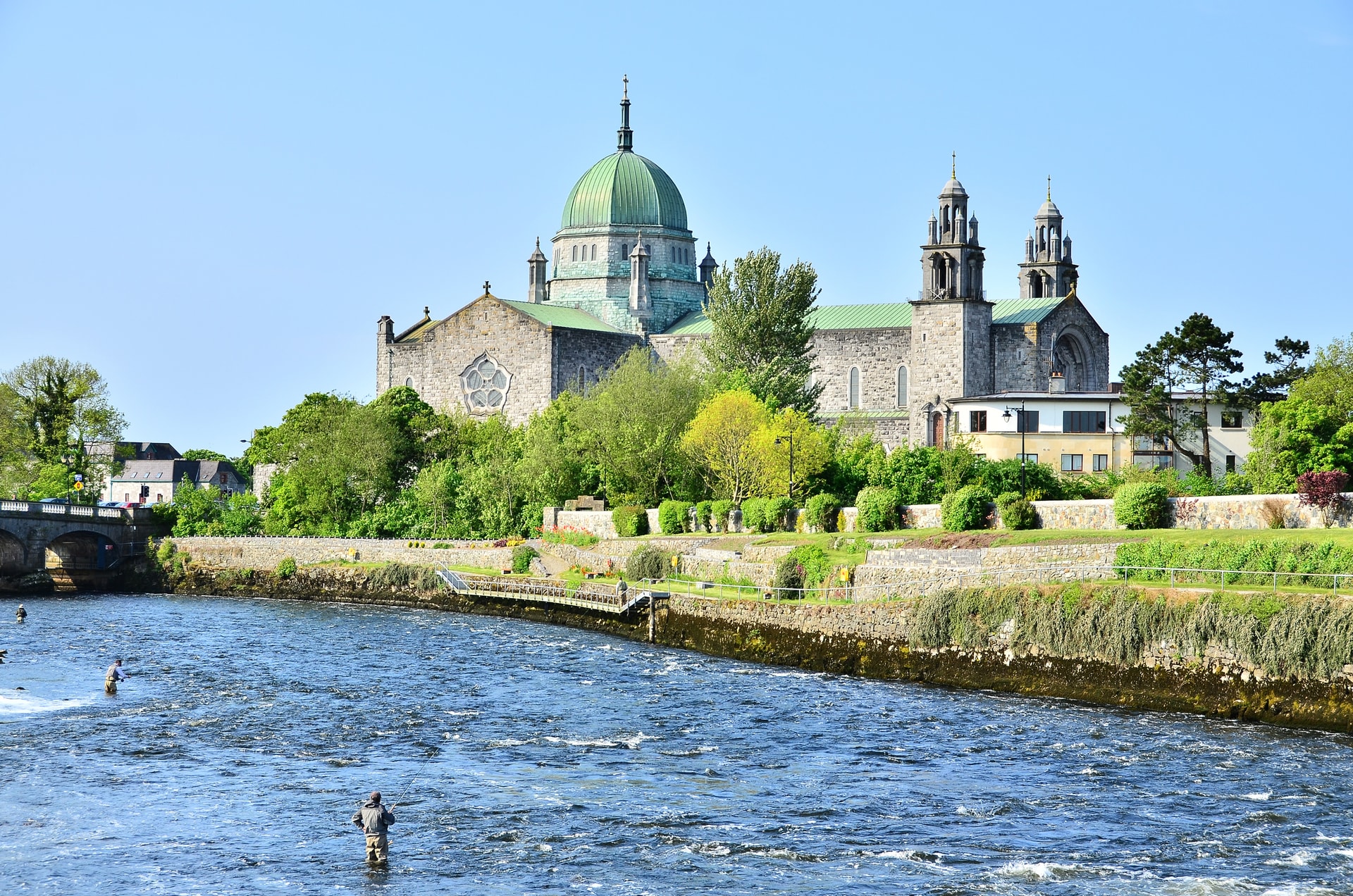 Galway Cathedral with green dome overlooking river where people are fly fishing under blue sky.