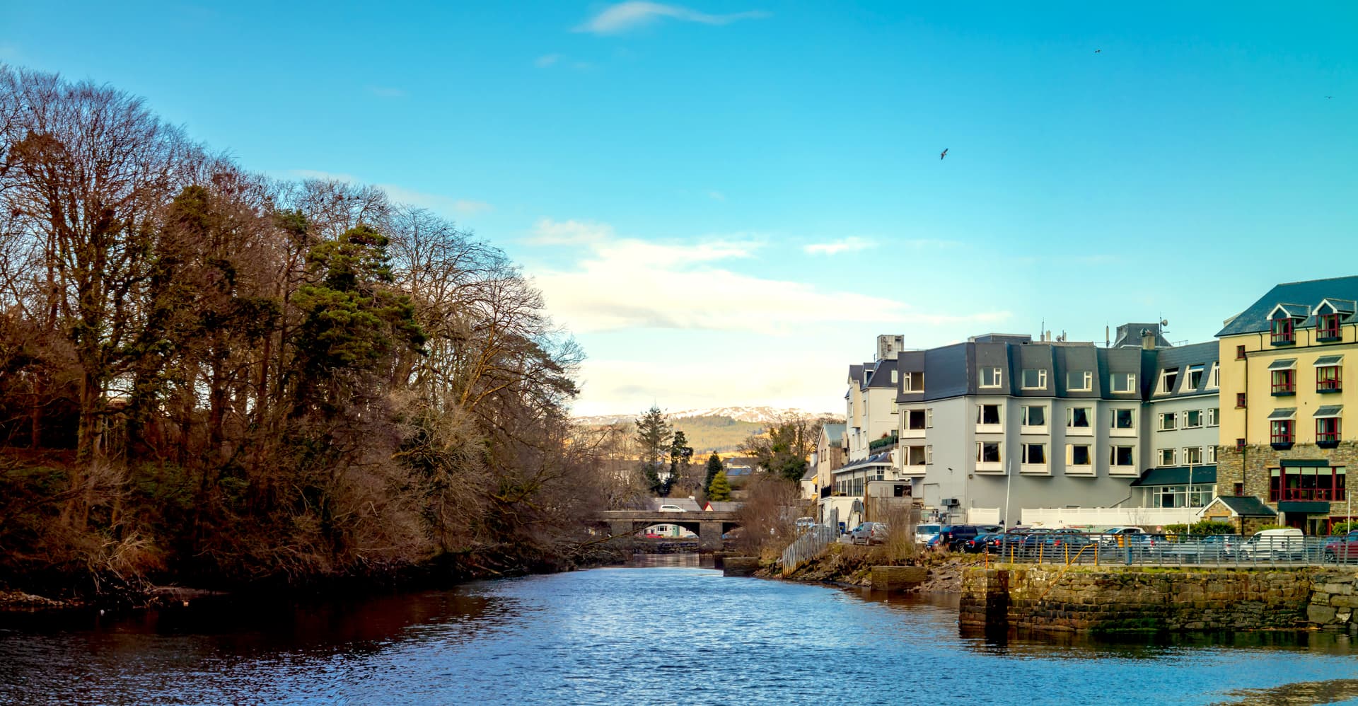 River flowing past buildings and bare trees in Donegal Town with snow-capped hills in the distance.