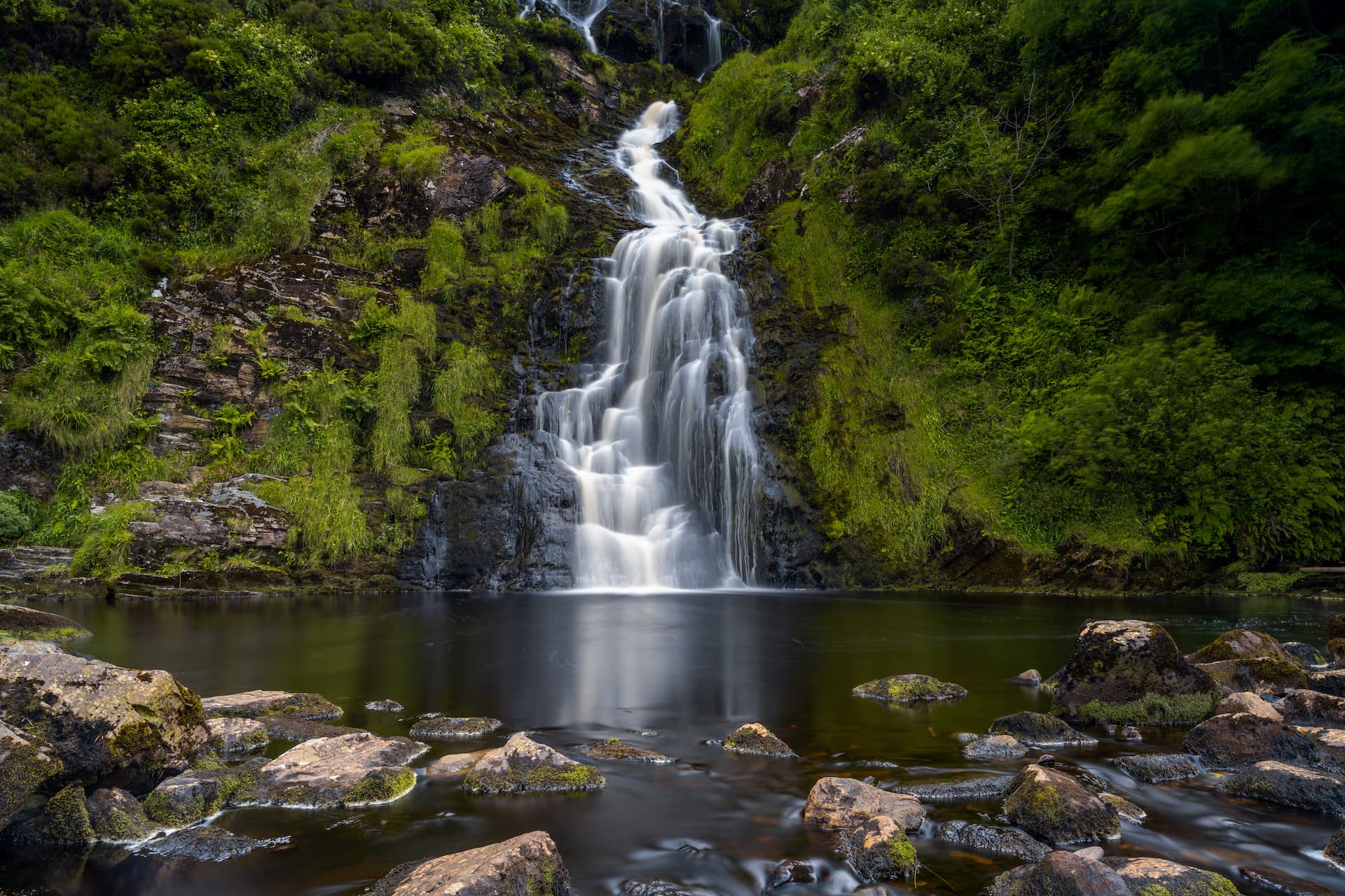 Assaranca Waterfall cascading down mossy rocks into a dark pool with foreground boulders.