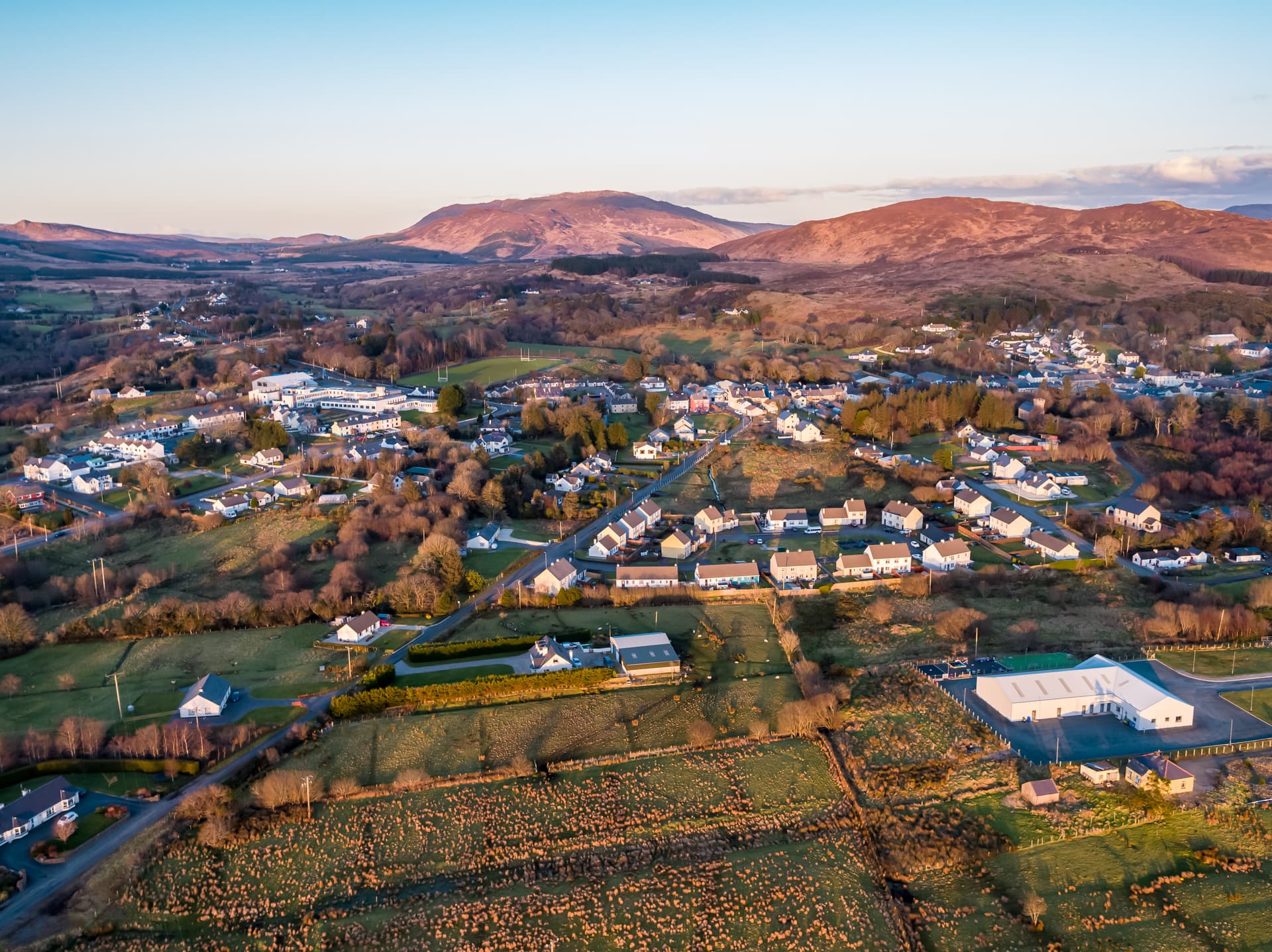 Aerial view of Glenties village nestled in green fields with brown mountains under a clear sky.