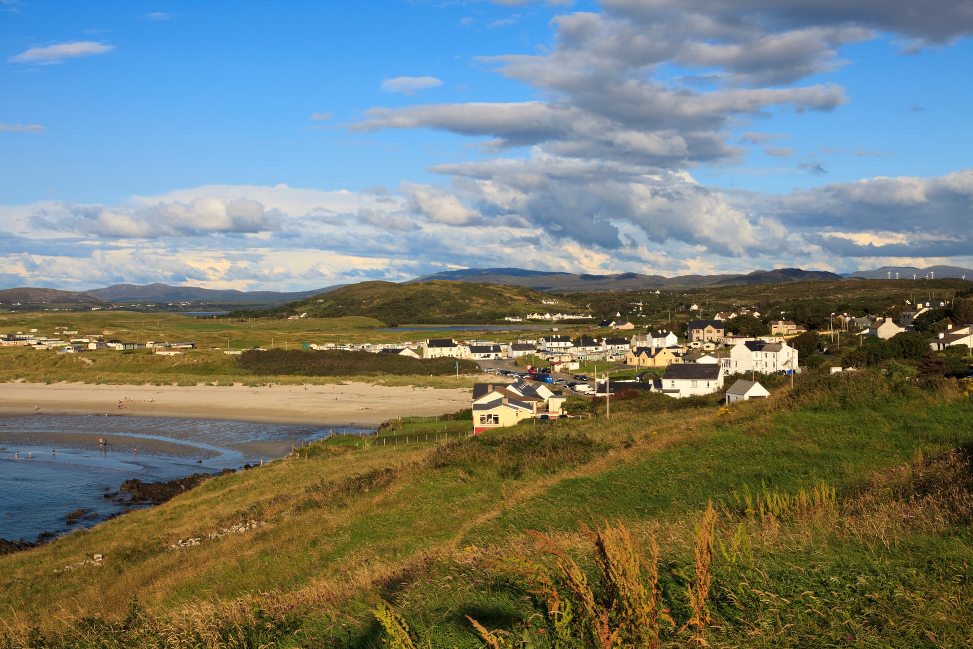 Coastal village near a sandy beach with green hills and mountains under a cloudy blue sky.