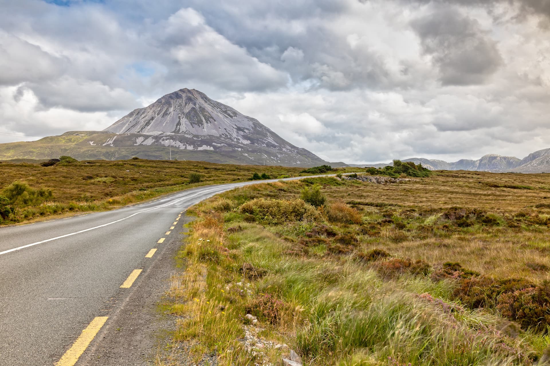 Winding road through grassy moorland toward a large, rocky mountain under a cloudy sky.