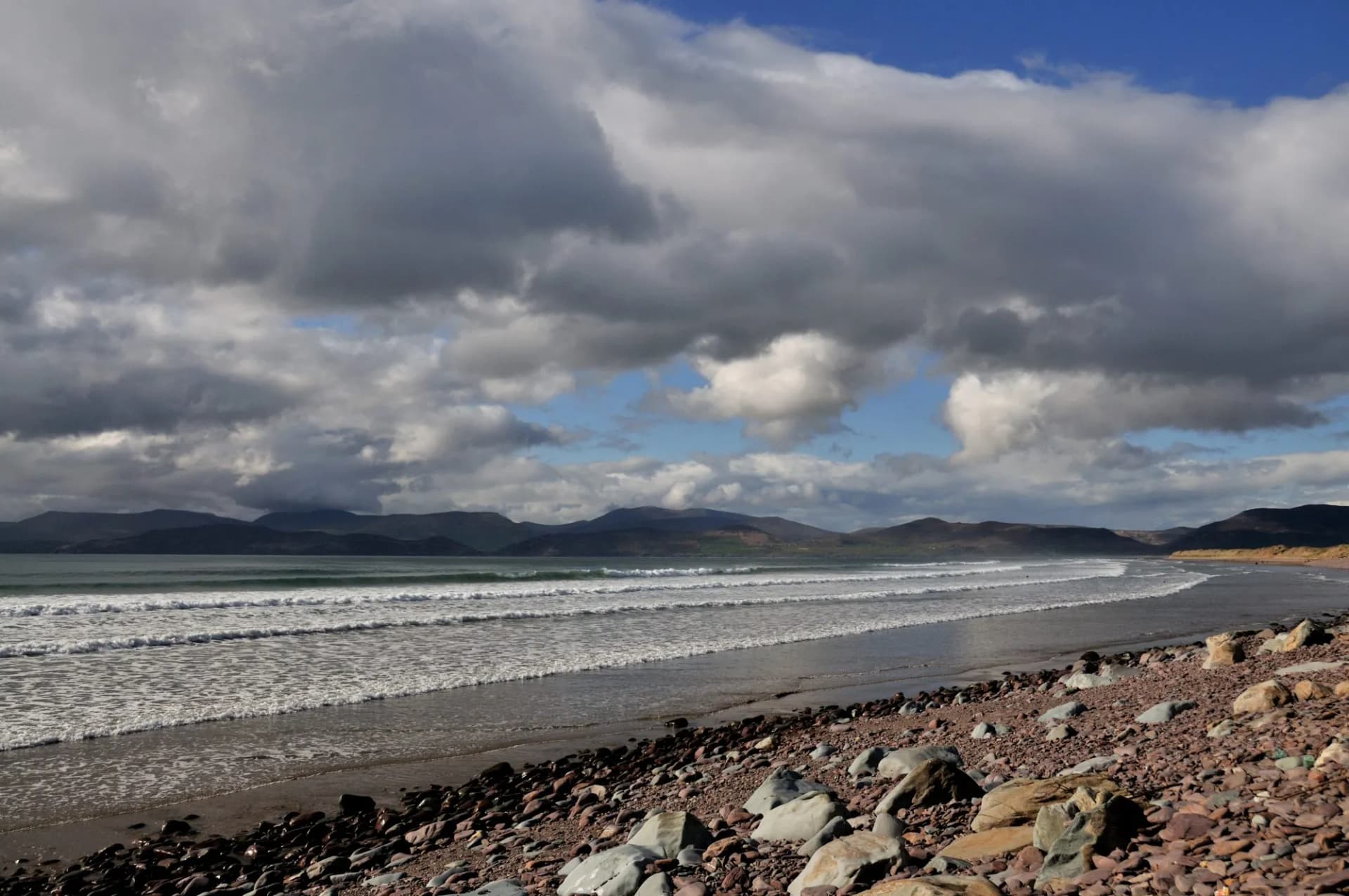 Rocky beach shoreline with waves rolling toward dark mountains under a dramatic cloudy sky