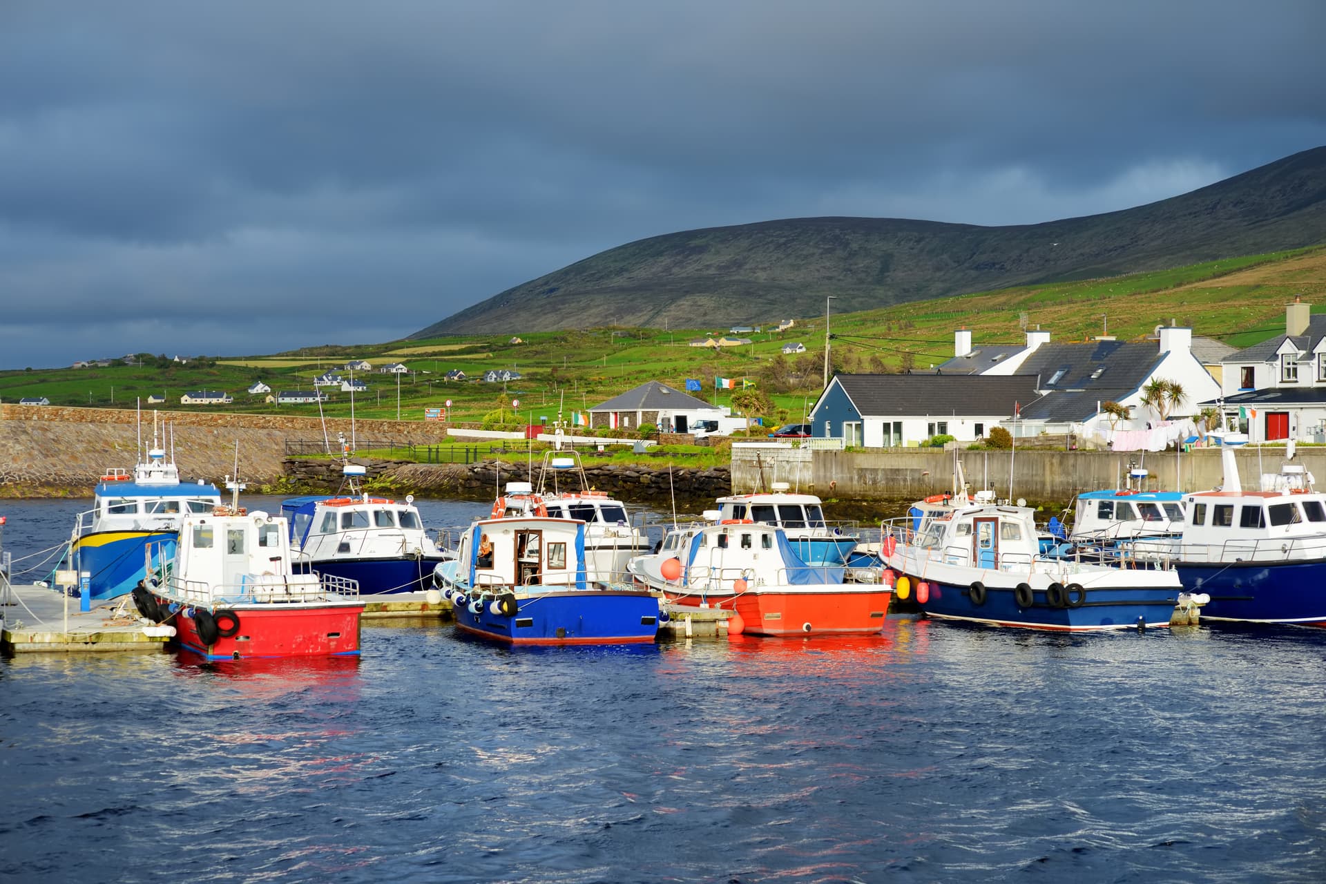Boats docked in Portmagee harbor with green hills and white houses under dark clouds.