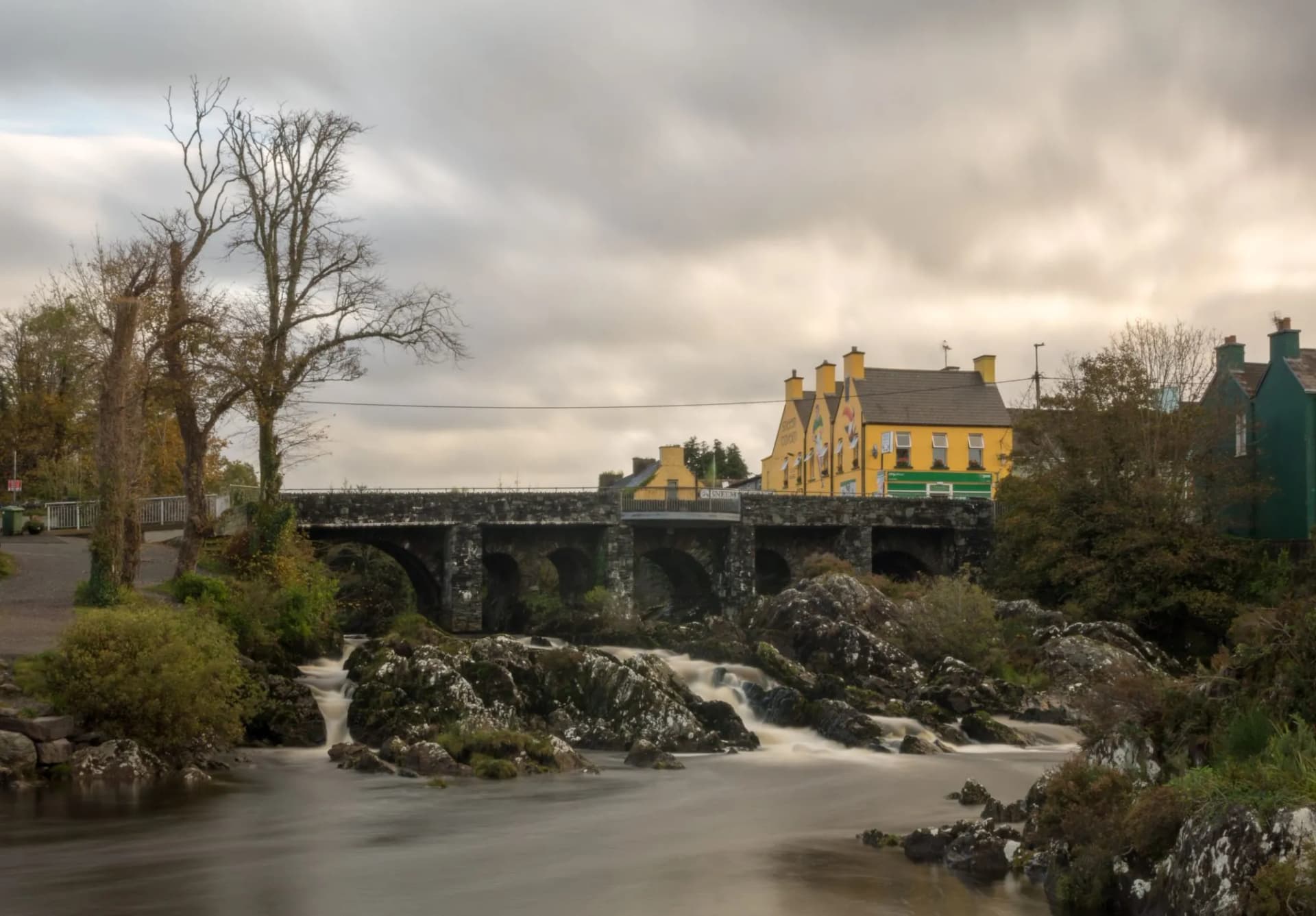 Stone arched bridge over rushing river rapids near colorful buildings in Village Sneem