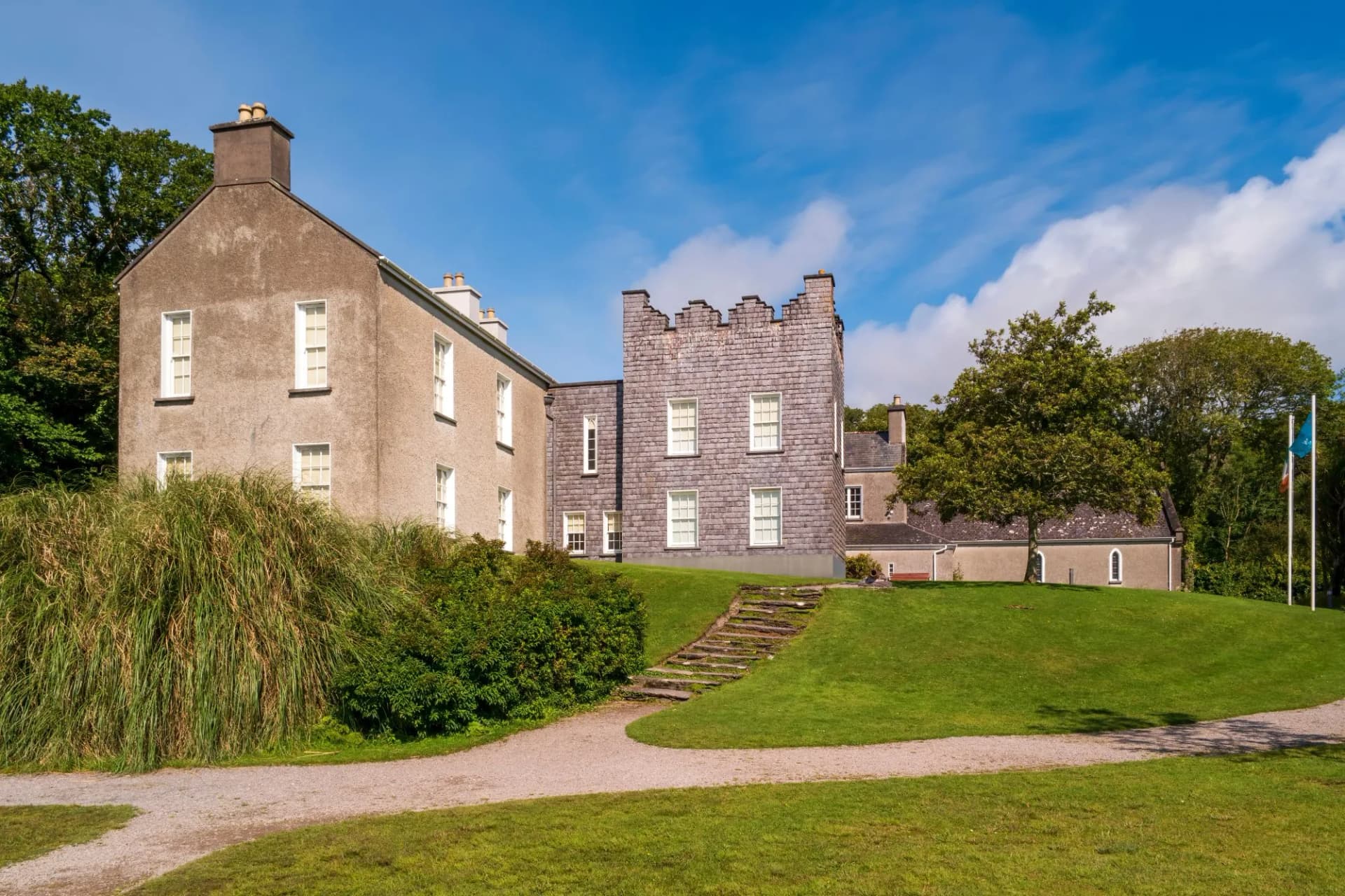 Derrynane House with crenellated tower section on a sunny day with green lawn and gravel path.