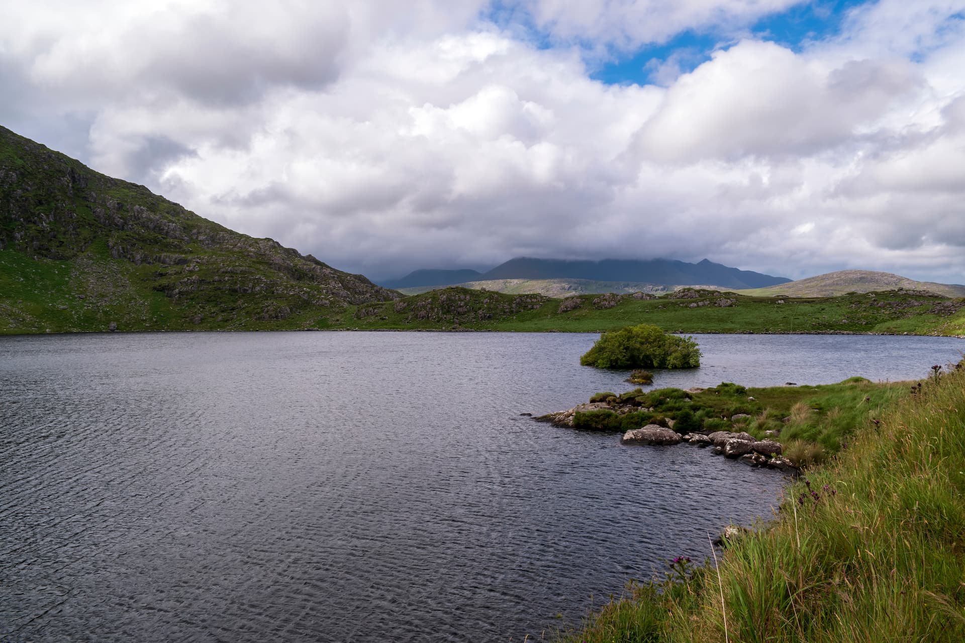 Dark water of Lough Barfinnihy with green, rocky mountains under a cloudy sky.