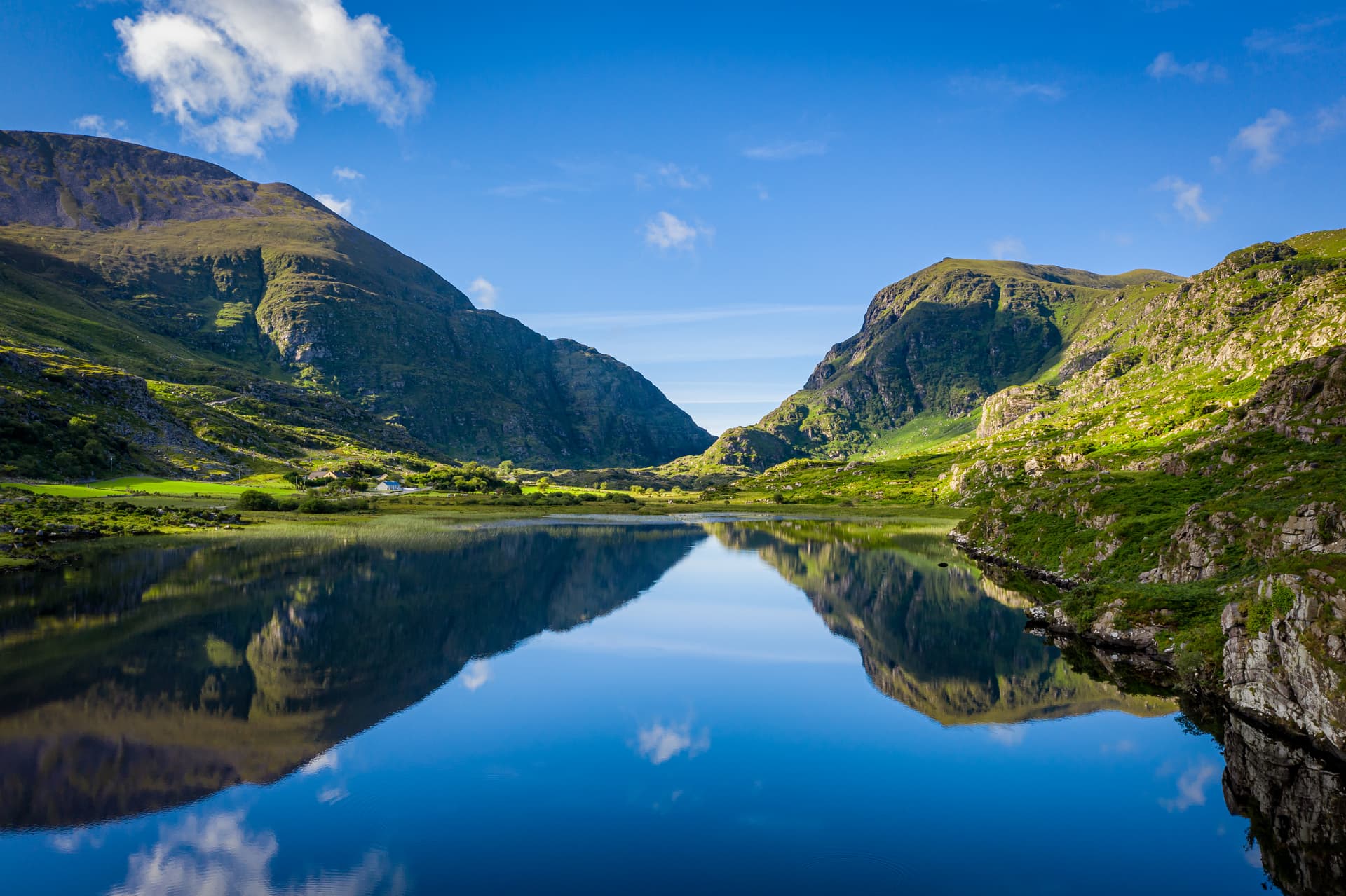 Gap of Dunloe lake reflecting steep green mountains under a bright blue sky with white clouds.